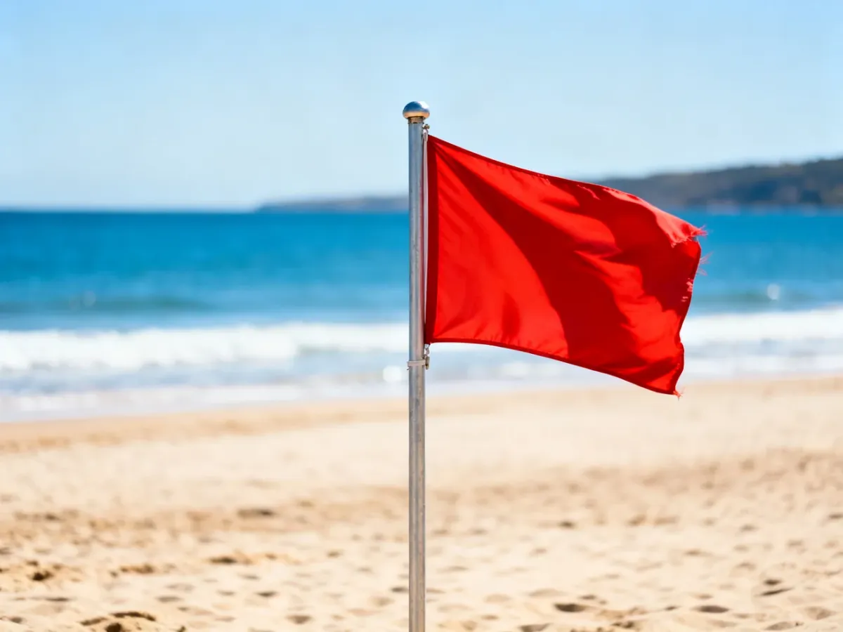 Czerwona flaga powiewająca na maszcie na piaszczystej plaży, oznaczająca zakaz kąpieli w morzu.